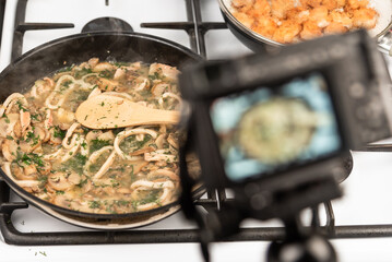 Shooting on the camera for food blog cooking creamy sauce with squid and mushrooms. Sliced squid rings and champignons, with dill and parsley stewed with cream in a pan