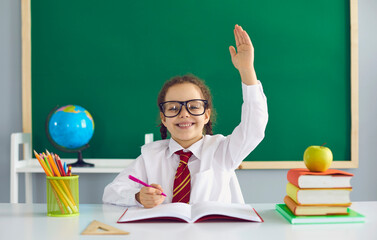 First day at school. A happy schoolgirl with glasses smiling raised her hand up while sitting at a table in the classroom against the background of the school blackboard.
