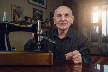 happy elderly bald tailor holds glasses sitting by black vintage sewing machine