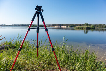 Camera red tripod standing in green grass near lake water natural landscape on clear sunny bright day