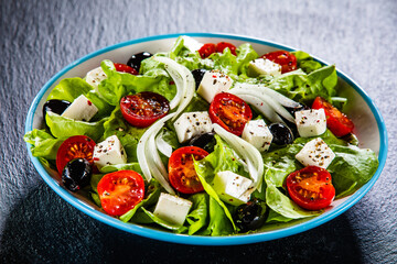 Fresh Greek salad - feta cheese, cherry tomatoes, lettuce, black olives and onion on black stone board
