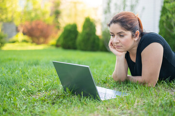A woman lies on her stomach on the lawn at the cottage and uses on a laptop. A focused female freelancer works remotely outdoors. The girl is studying on the computer.
