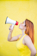 A woman in a dress with glasses and earrings stands in profile on a yellow background and shouts in a megaphone. Portrait of a girl holding a loudspeaker. Lady yells at a sound amplification device.
