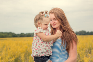Fototapeta na wymiar A little beautiful happy girl in her mother's arms. In everyday clothes. Smiling in a yellow field. With ponytails on their heads. Horizontal photo. Cloudy weather. Summer.