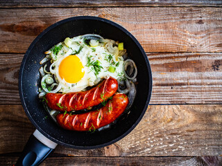 Fried sausages with egg and onion in frying pan on wooden table