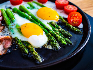 Preparing one pen breakfast. Sunny side up eggs with green asparagus, fried bacon and cherry tomatoes 
