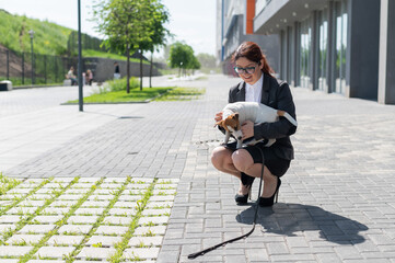 Business woman in a suit and high heels hugs a puppy of Jack Russell Terrier on the street. Little smart dog plays with the owner in the fresh air. Love for the pet.