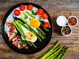 One pen breakfast. Sunny side up eggs with green asparagus, fried bacon and cherry tomatoes on black plate on wooden table
