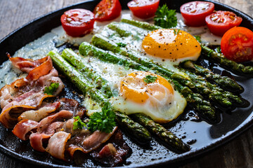 One pen breakfast. Sunny side up eggs with green asparagus, fried bacon and cherry tomatoes on black plate on wooden table
