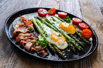 One pen breakfast. Sunny side up eggs with green asparagus, fried bacon and cherry tomatoes on black plate on wooden table
