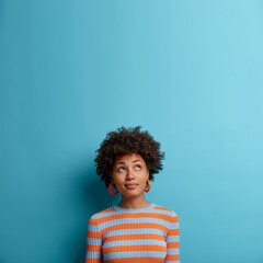 Vertical shot of cute dark skinned woman with Afro hairstyle, looks above with pondering expression, notices something interesting, blank space on blue background. Face expressions and emotions - obrazy, fototapety, plakaty