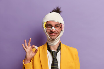 Funny glad man with shiner and broken nose, wears bandage on head, makes okay gesture, assures that...