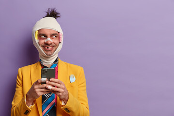 Happy pleased injured man holds modern smart phone, types messages and scrolls social networks,...