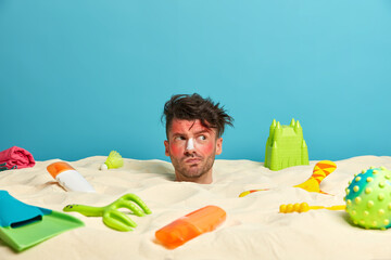 Young man gets suntan at beach, buried in sand, has sunburn face skin, applies sunnscreen on nose,...