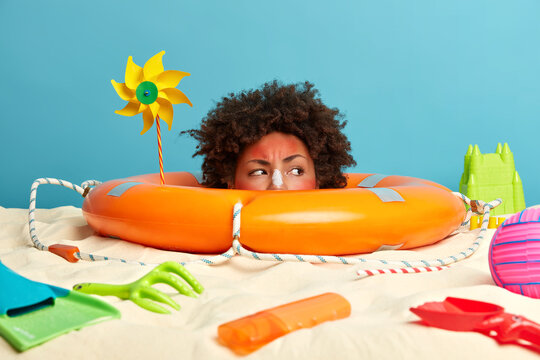 Recreation and summer vacation concept. African American woman with displeased expression, sunbathes during hot day, sticks out head from lifebuoy, poses at beach, got suburn, applies sunscreen lotion