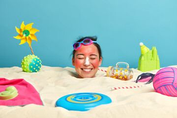 Happy ethnic lady with applied sunblock cream, sunbathes on sandy beach, has fun and good mood,...