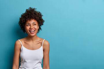 Portrait of optimistic dreamy woman with Afro hair, imagines something positive, looks somewhere and dreams, wears casual vest, isolated on blue background, blank copy space for your advertisement