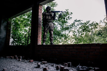 Military in camouflage standing with a rifle in the doorway of an abandoned building aiming at the...