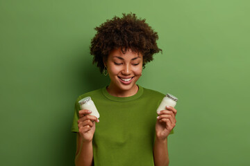 Portrait of happy woman holds two bottles of healthy yoghurt, chooses dairy product for breakfast, keeps to diet, poses indoor against green background. People, lifestyle and nutrition concept