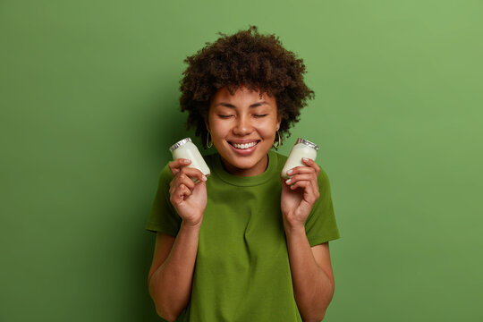 Happy woman with curly hair holds two jars of vegan plant based yogurt, smiles pleasantly with closed eyes, has proper healthy nutrition, wears green t shirt, poses indoor. Female with dairy product