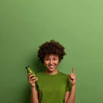 Smiling glad dark skinned curly girl points above with fore finger, holds glass bottle of fresh vege
