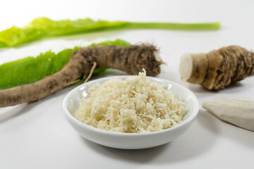 Grated horseradish in a small bowl, fresh roots and leaves blurry in the light gray background, selected focus