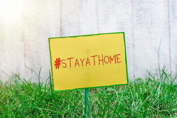 Handwriting text writing Hashtag Stay at home. Conceptual photo a trending label in social media related to the coronvirus outbreak Plain empty paper attached to a stick and placed in the green grassy