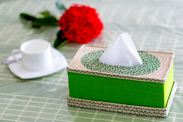 Green Handmade fiber Napkin box on the dining table with a red flower and a white tea-cup. Tissue paper box on restaurant.
