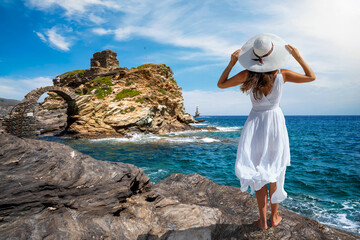 A tourist woman in a white dress stands at the coast of Andros island and enjoys the view to the old stone bridge leading to the ancient castle, Cyclades, Greece