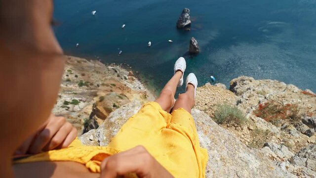 camera zooming out first person view of taned woman legs in yellow dress sitting on on cliff edge. amazing nature view of seascape with high limestone cliff over blue sea and clear deep blue sky