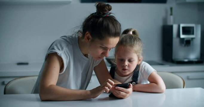 Mom and daughter sit at a table in the kitchen. Mom teaches a little daughter to use the phone. Close-up portrait of a woman