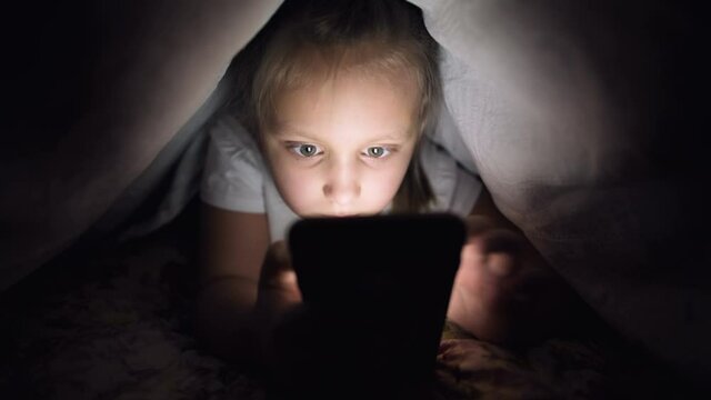 A keen little girl plays on a smartphone at night under a blanket. The child's departure from reality through mobile devices. Child uses secret phone