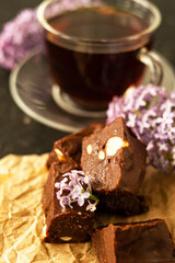 A piece of homemade chocolate cake on crumpled craft paper, with nuts on a black texture table. A cup of tea and lilac flowers decorate a delicious tea party.