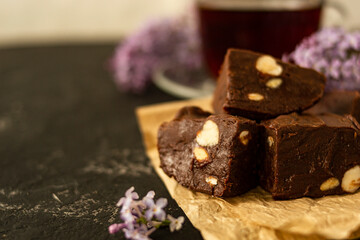 A piece of homemade chocolate cake on crumpled craft paper, with nuts on a black texture table. A cup of tea and lilac flowers decorate a delicious tea party.