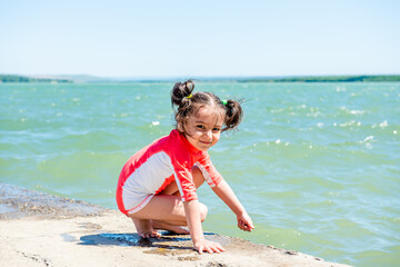 little cute girl in a pink bathing suit sits on a pier by the lake on a background of blue water. Place for text