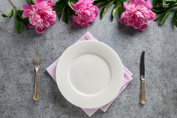 Festive table setting with pink peony flowers, white dishware, silverware on gray. Top view.