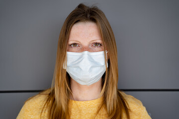 Caucasian woman wearing white disposable surgical face mask. Grey wall in background. Corona virus...