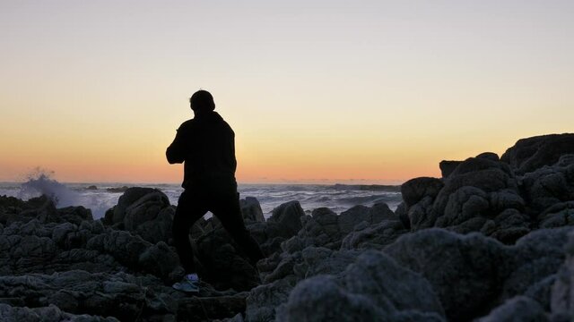 Men warrior monk practicing silhouette tai chi karate kung Fu on the rocky stones horizon at sunset or sunrise. Art of self-defense. Silhouette on a background of dramatic epic waves at pacific coast