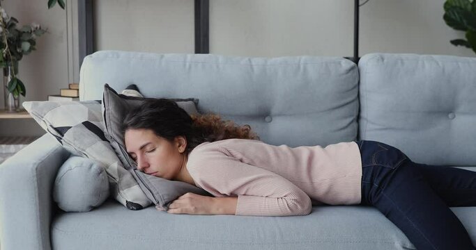 Unhappy young woman falling down on comfort couch at home, exhausted after hard workday. Tired beautiful lady feeling lack of motivation inspiration, sleeping on sofa indoors, depression concept.
