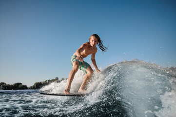 young active man rides on surfboard on wave against clear blue sky.