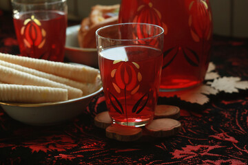 Jug with red compote, glasses, puff with tropical fruits, banana wafer rolls, a bouquet of yellow mimosa in a vintage vase, in the kitchen on the table covered with a red brocade tablecloth.