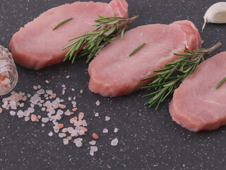 
three raw steaks with sprinkled pink salt and two sprigs of fresh rosemary on a black cutting board, closeup side view.