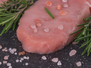 
one raw steak with sprigs of fresh rosemary on a black cutting board, closeup side view.