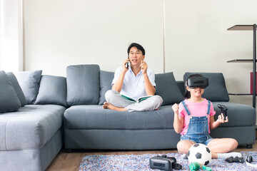daughter sitting on carpet in living room and holding controller during playing game via virtual reality headset while asian father calling with smartphone on sofa.