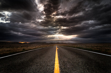 Lonely asphalt highway heading into a sunset with a cloudy sky and sun rays. There is a yellow...