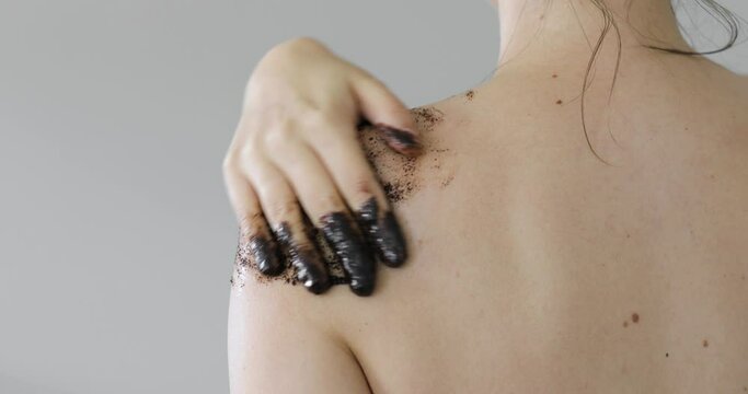Woman is applying black eco organic natural coffee scrub on shoulder on grey background, closeup back view. Beauty procedure, spa, wellness and healthy skin care concept. Scrubbing skin.