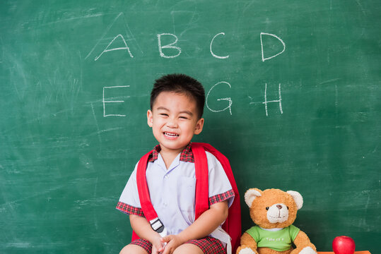 Back to School. Happy Asian funny cute little child boy kindergarten preschool in student uniform with school bag, book sit with teddy bear on green school blackboard, First time to school education