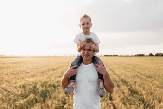 Portrait of a happy father gives his son a contrailer to ride on his shoulders and look up. Cute boy with dad plays outdoors wearing white t-shirts.