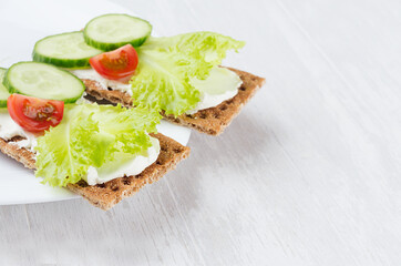 Fototapeta na wymiar Fresh summer healthy appetizer of whole grain rye crisps breads with vegetables - green salad, cucumber, tomato, cream cheese on white wood board, closeup, copy space.