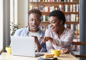 Joyful black couple watching funny movie together on laptop at coffee shop